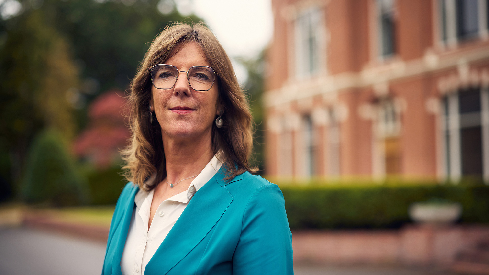 Portretfotografie Zuidlaren – Vrouw In Blauw-Groen Pak Kijkt Krachtig Naar Camera Met Gebouw En Groen Op Achtergrond